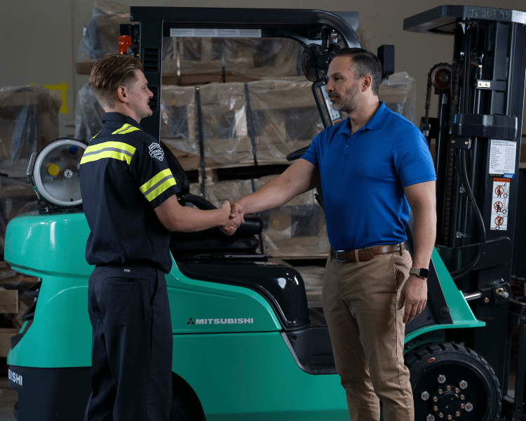 An Equipment Depot forklift technician shaking hands with a customer in front of a Mitsubishi forklift inside a warehouse.